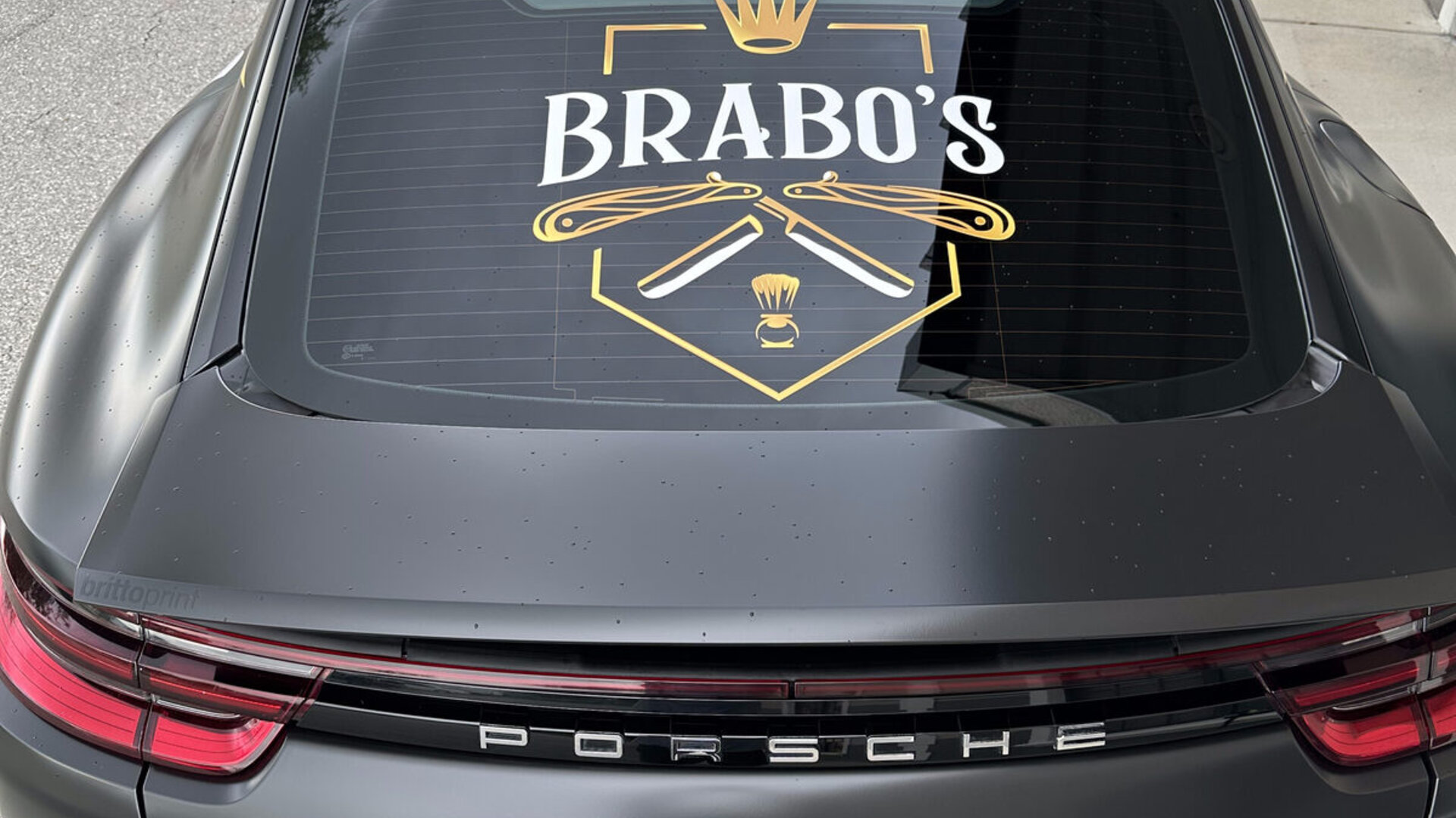 Porsche Panamera wrapped in Avery Satin Black with Brabo's Barbershop glossy printed crest and lightning bolts on the hood — finished install photographed at Brittoprint, Lehigh Acres / Fort Myers, FL. © 2026 Brittoprint.
